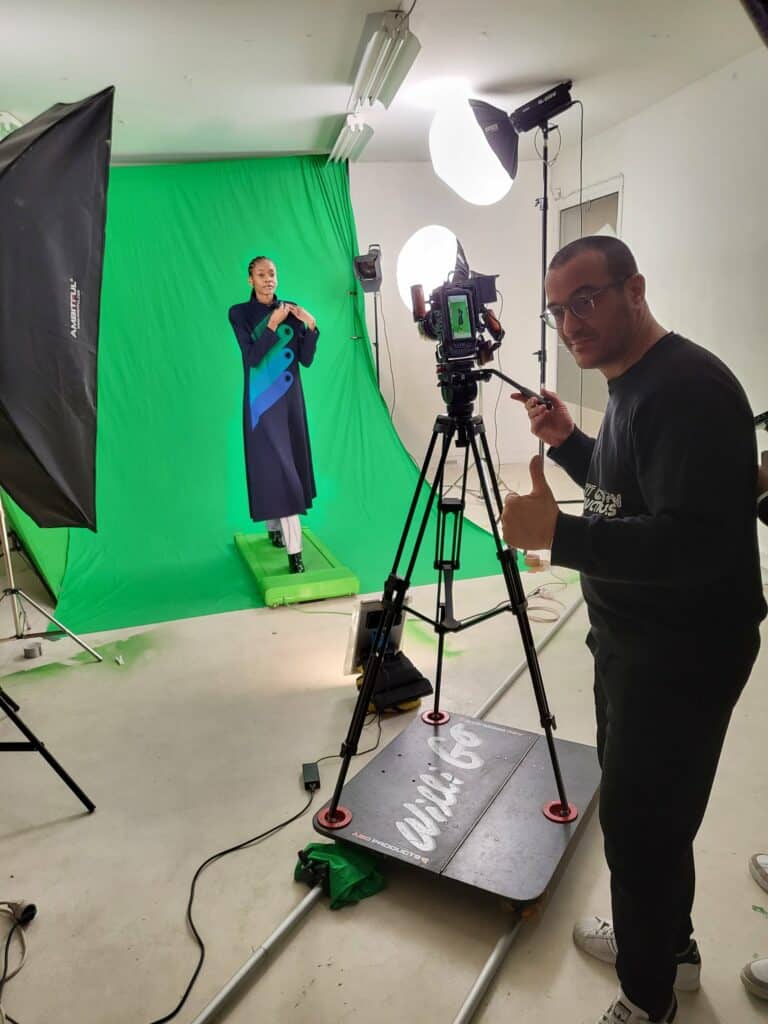Tournage sur fond vert pour Pierre Cardin : un mannequin en manteau bleu nuit marche sur un tapis roulant, David Lévy règle la caméra et la lumière
