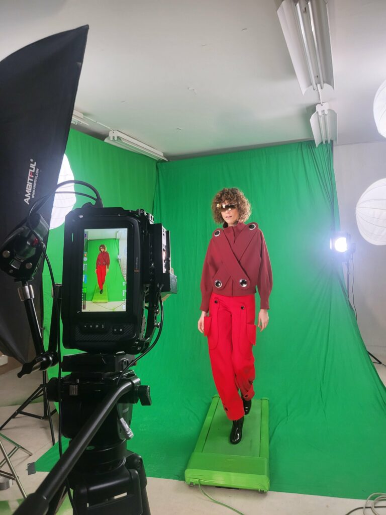 Mannequin en veste bordeaux et pantalon rouge, lunettes futuristes, filmée sur fond vert devant le moniteur de la caméra